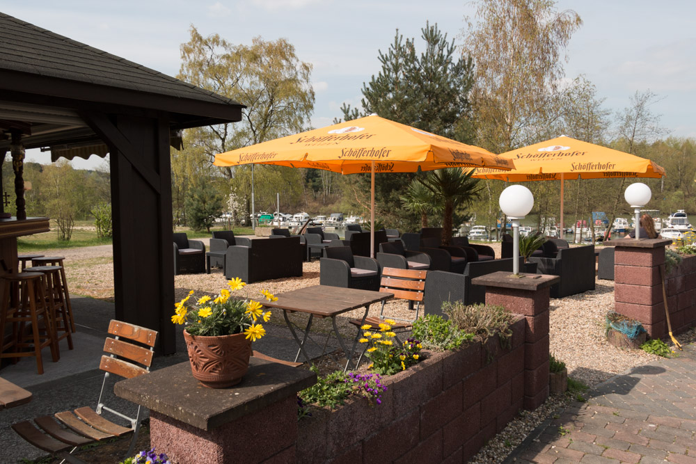 Terrassenbereich mit schwarzen Stühlen, Holztischen, gelben Sonnenschirmen, Topfblumen und einer Bar mit Blick auf einen Jachthafen mit Booten im Hintergrund.