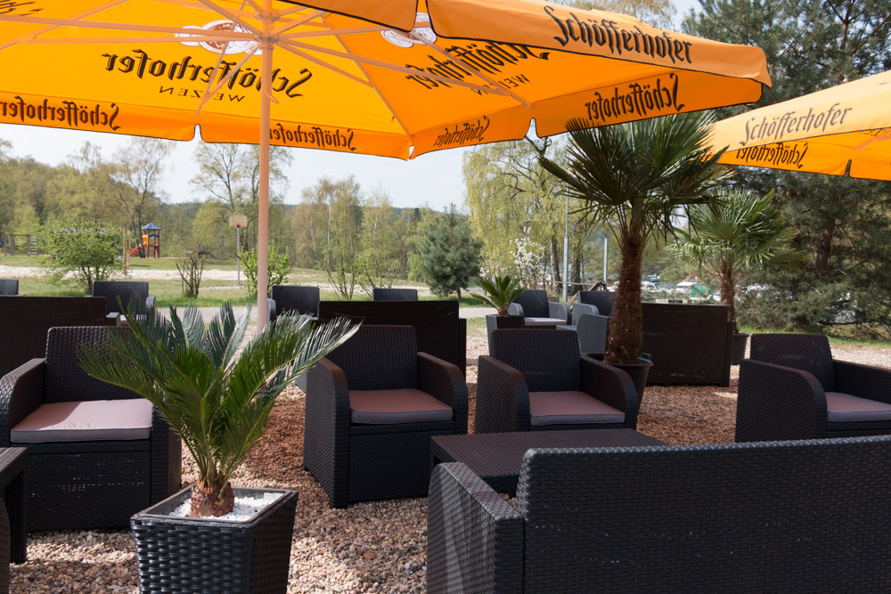 Sitzbereich im Freien mit schwarzen Korbstühlen und -tischen, gelben Markenschirmen und getopften Palmen auf einer Kiesfläche, umgeben von Bäumen und Grünpflanzen.