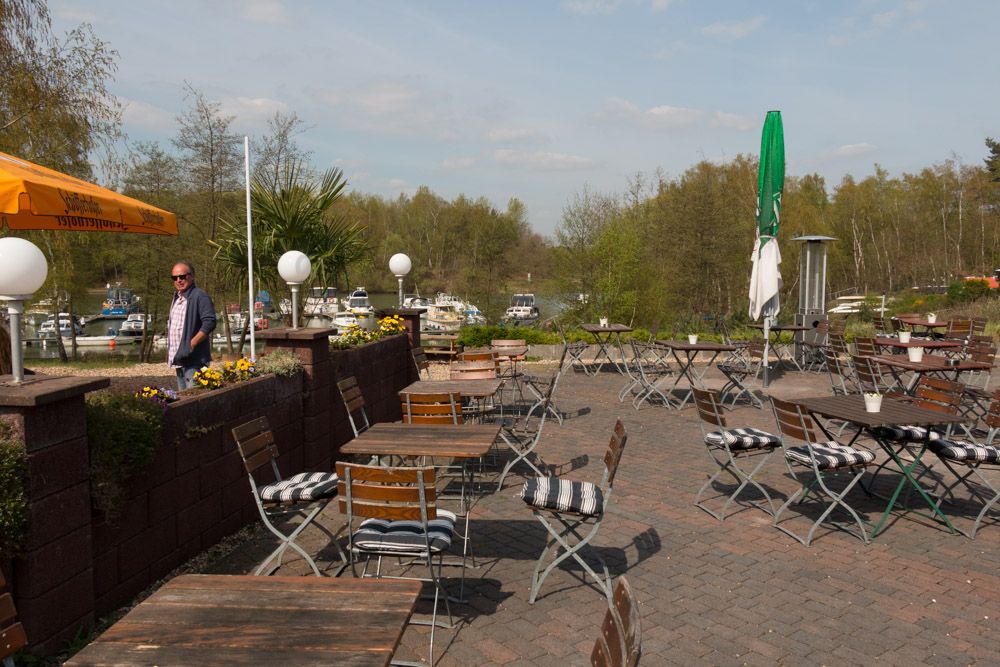 Café-Sitzgruppe im Freien mit leeren Tischen und Stühlen, ein Mann steht an einer niedrigen Mauer, Boote und Bäume im Hintergrund unter einem teilweise bewölkten Himmel.