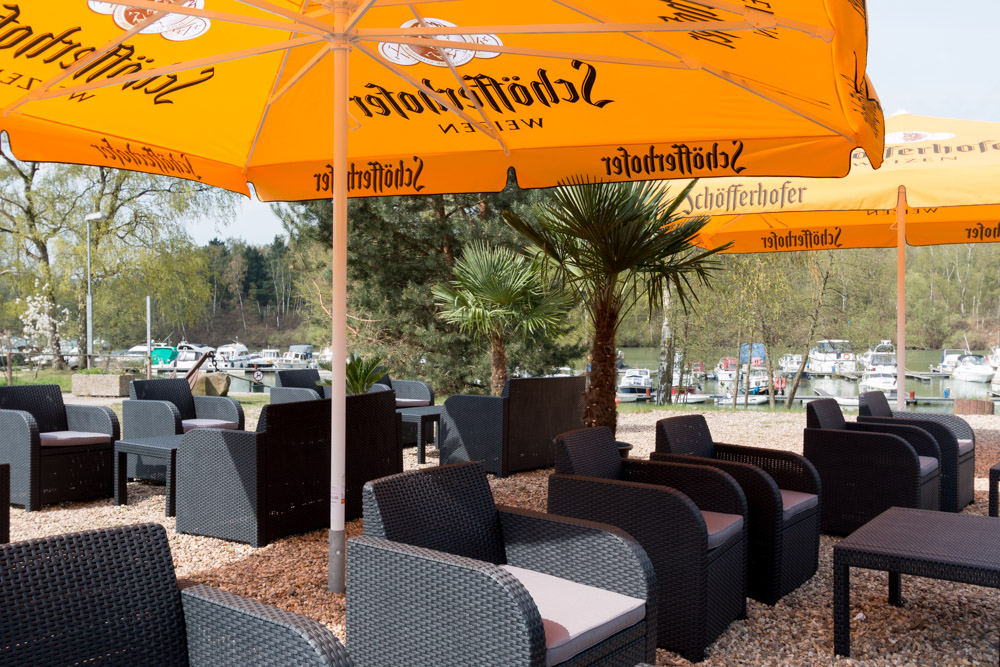 Sitzbereich im Freien mit schwarzen Korbstühlen und Tischen unter großen gelben Sonnenschirmen, umgeben von Bäumen und mit Blick auf einen Yachthafen mit Booten.