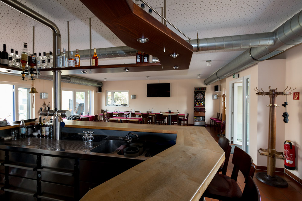 Eine moderne Bar mit Holztresen, Metallbeschlägen und Flaschenregalen blickt auf einen Sitzbereich mit Tischen, Stühlen, einer Dartscheibe und einem an der Wand montierten Fernseher.