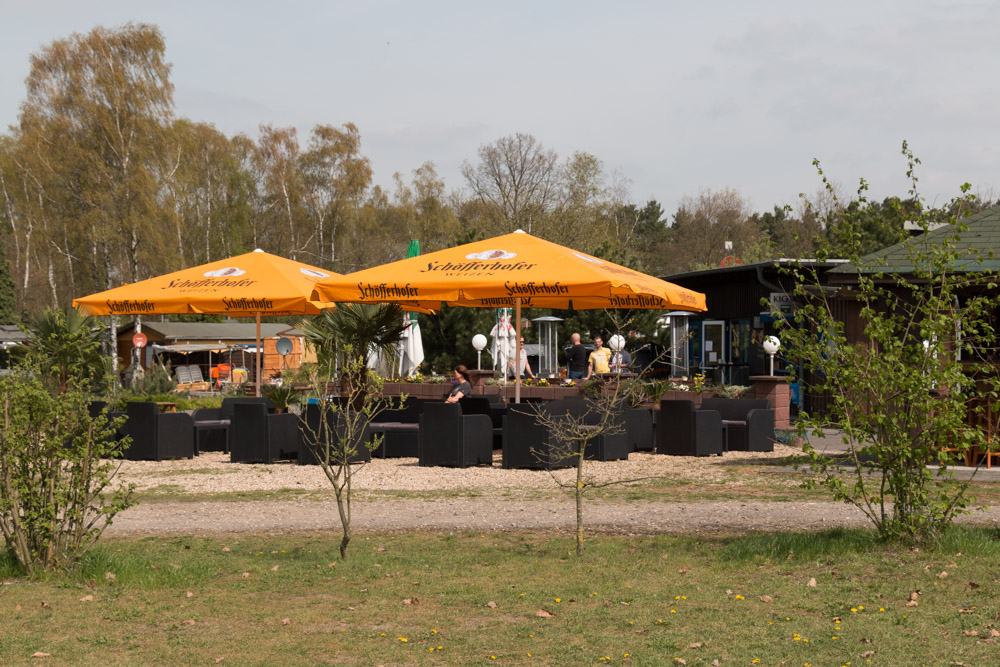Sitzbereich im Freien eines Cafés oder Restaurants mit großen gelben Sonnenschirmen, schwarzen Möbeln und einigen sitzenden Personen; umgeben von Grün und Bäumen.