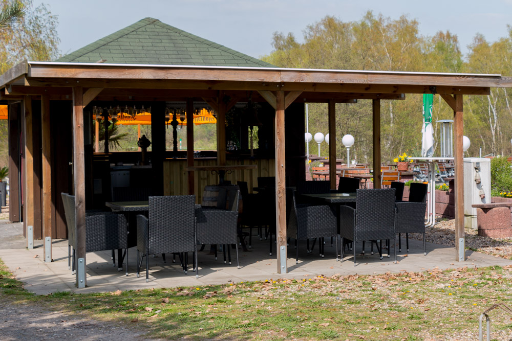 Holzpavillon mit grünem Dach, in dem mehrere schwarze Tische und Stühle auf einer gepflasterten Terrasse stehen, umgeben von Bäumen und Herbstlaub.