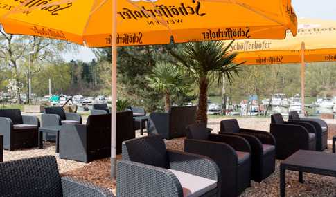 Sitzbereich im Freien mit schwarzen Korbstühlen und Tischen unter großen orangefarbenen Sonnenschirmen, umgeben von Palmen und einem Yachthafen mit Booten im Hintergrund.