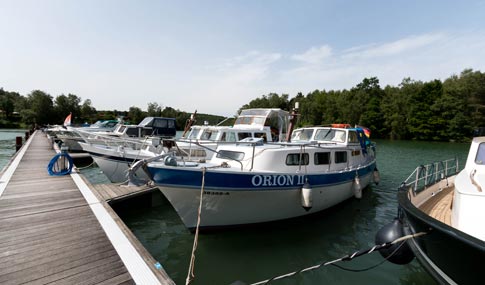 Mehrere Motorboote, darunter eines mit dem Namen ORION II, liegen an einem Holzsteg auf einem ruhigen See mit Bäumen im Hintergrund.