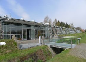 Ein modernes Glasgebäude mit schrägem Dach und Metallgerüst, umgeben von Gras und Bäumen, mit einer kleinen Fußgängerbrücke über einen Graben davor.