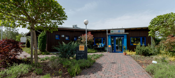 Ein einstöckiges Gebäude mit blauen Fensterrahmen und einem blauen Eingang, umgeben von Bäumen und einem angelegten Garten mit einem gemauerten Weg, der zur Tür führt.