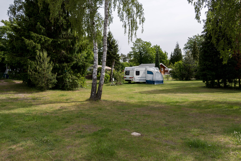Ein weißes Wohnmobil mit Markise steht auf einer von Bäumen umgebenen Wiese, und im Hintergrund ist ein kleines rotes Haus zu sehen.