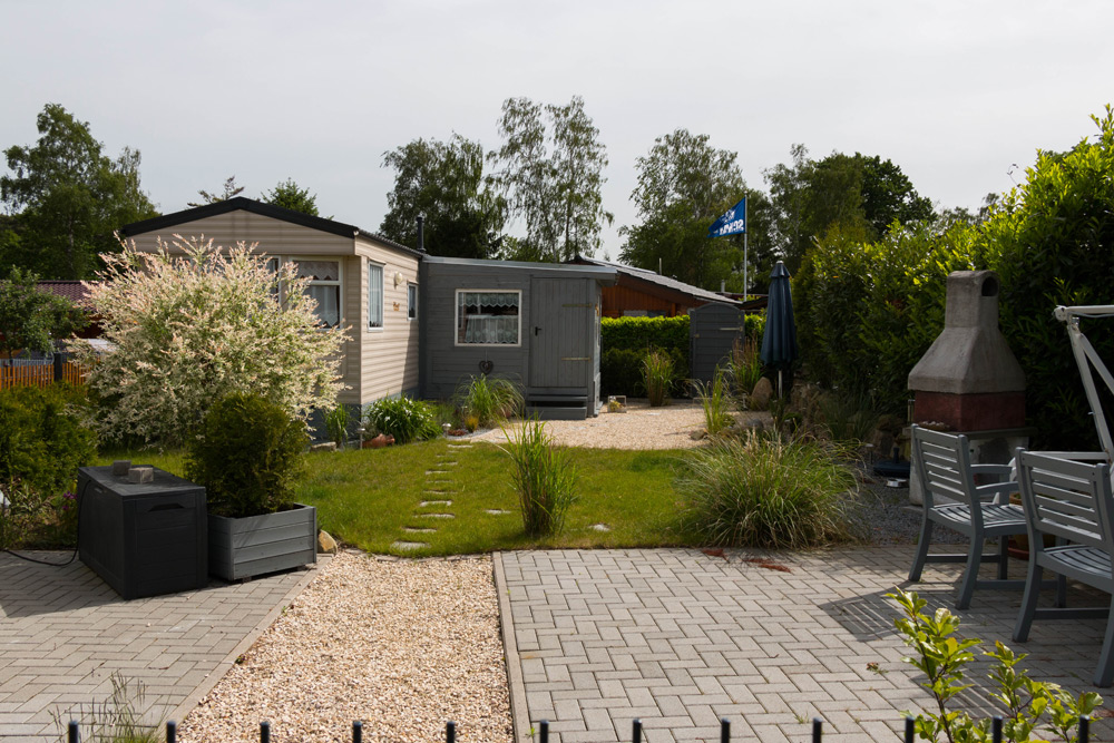 Ein kleiner Hinterhof mit einer gepflasterten Terrasse, einem Essplatz im Freien, einem Grillkamin, einem Schuppen und grünem Rasen, der von Sträuchern und Bäumen gesäumt wird.