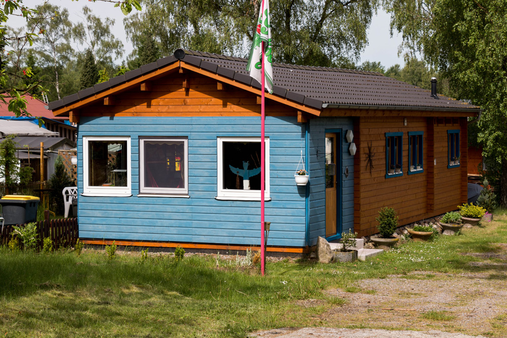 Einstöckiges Holzhaus mit blauem und braunem Äußeren, drei Fenstern an der Vorderseite, einer kleinen Veranda und einem Fahnenmast im Vorgarten, umgeben von Grünflächen.