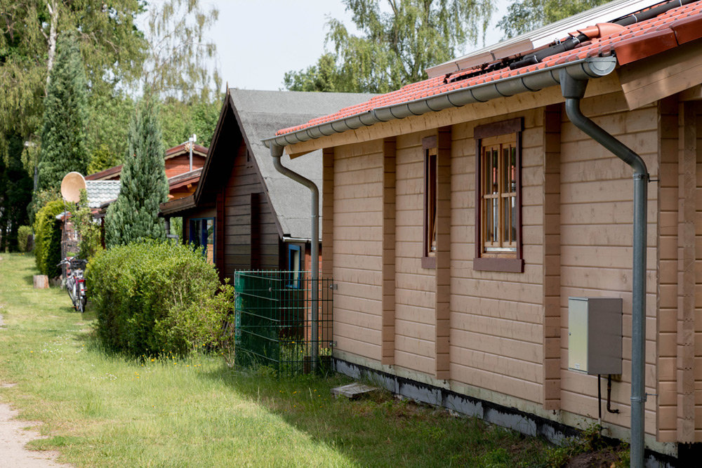 Eine Reihe von kleinen Holzhäusern mit roten Dächern und Fenstern, umgeben von Gras, Bäumen und Sträuchern an einem sonnigen Tag.