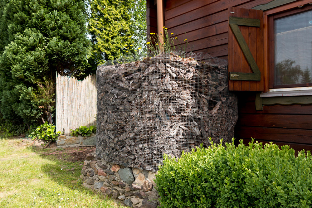 Eine mit Rindenmulch gefüllte Gabionenwand steht neben einem Holzhaus, das von grünen Sträuchern und einem Zaun umgeben ist, mit Bäumen im Hintergrund.