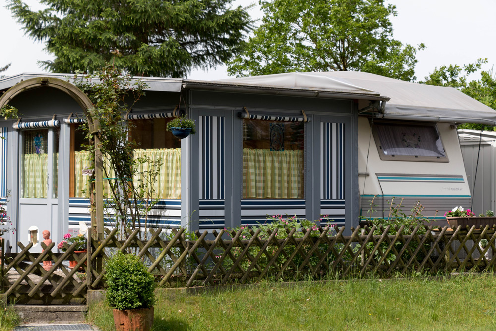 Ein stationärer Wohnwagen mit angebautem Vorzelt, blau-weiß gestreiften Paneelen, Gitterzaun und Topfpflanzen auf einer Wiese mit Bäumen im Hintergrund.