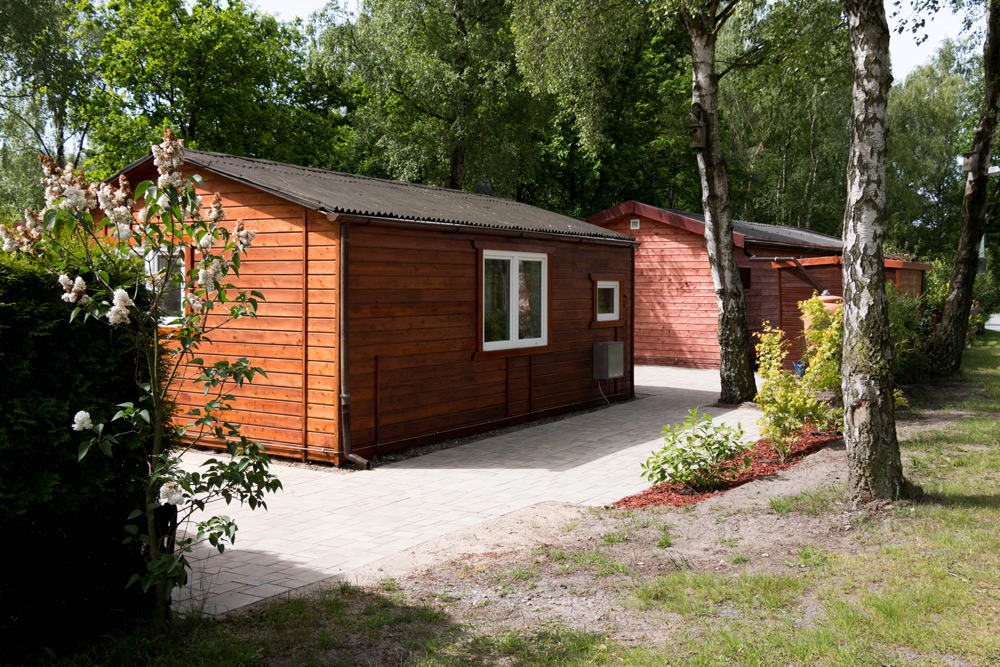 Zwei kleine Holzhütten mit Fenstern und Türen liegen an einem gepflasterten Weg, umgeben von Bäumen und Grün in einem bewaldeten Außenbereich.