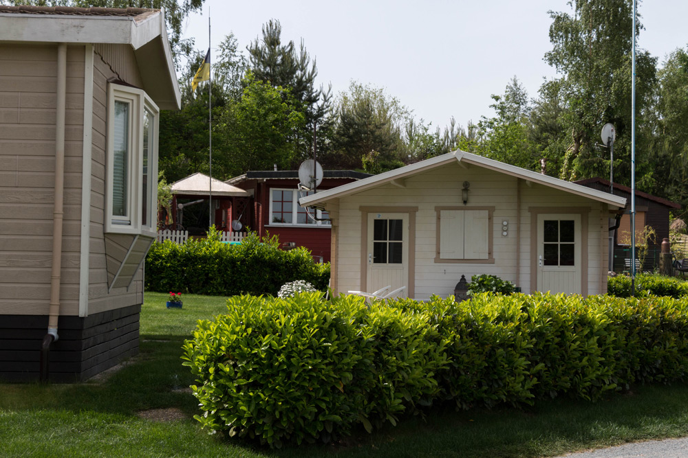 Eine kleine weiße Hütte mit Fensterläden steht hinter einer grünen Hecke, im Hintergrund sind weitere Häuser und Bäume zu sehen.