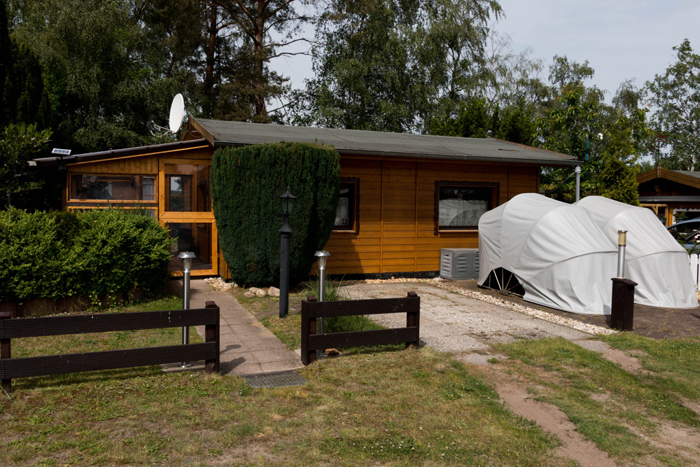 Ein kleines Holzhaus mit grauem Dach, Satellitenschüssel und überdachten Fahrzeugen in der Einfahrt, umgeben von Grünflächen und einem niedrigen Zaun davor.