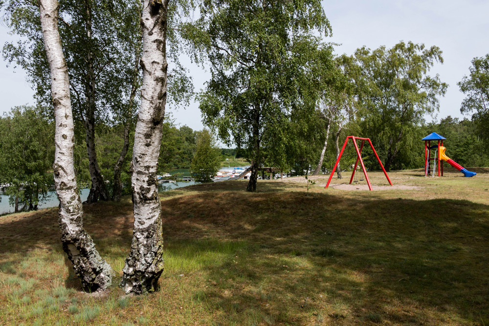 Spielplatz mit einer roten Schaukel und einer blau-gelben Rutsche zwischen Bäumen, mit Booten auf dem Wasser im Hintergrund.