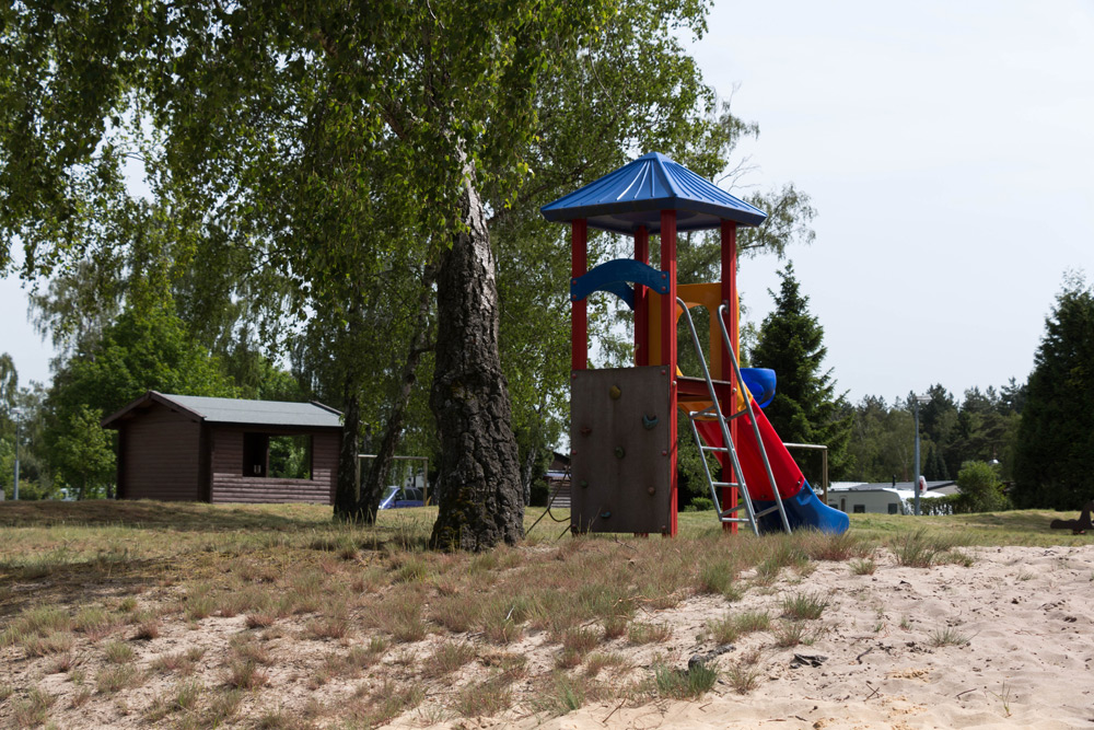 Ein Spielplatz mit einem blauen und roten Kletterturm, einer Rutsche und einer Leiter liegt auf sandigem Boden in der Nähe einer kleinen Holzhütte und grüner Bäume.