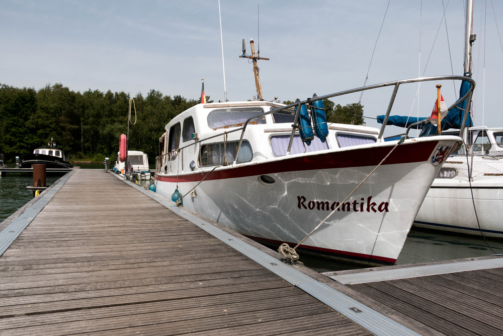 Ein weißes Boot mit dem Namen Romantika liegt an einem Holzsteg, im Hintergrund sind Bäume und andere Boote zu sehen.