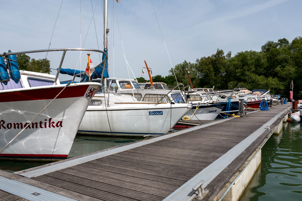 Mehrere festgemachte Boote liegen aufgereiht an einem hölzernen Steg an einem ruhigen Gewässer, mit Bäumen im Hintergrund.