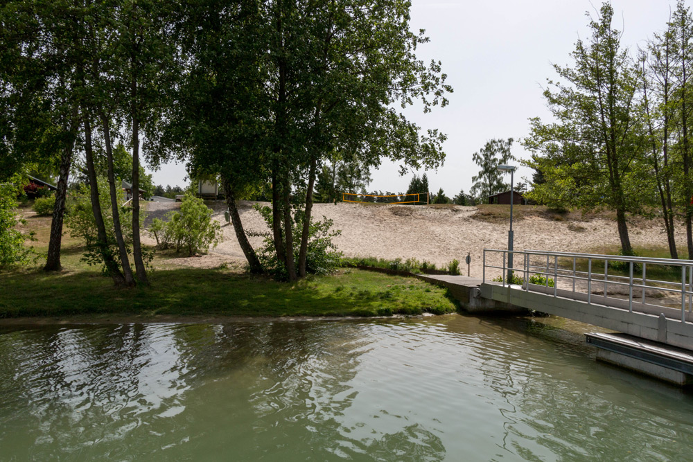 Eine kleine Fußgängerbrücke führt über das Wasser zu einer sandigen Fläche mit Bäumen und Grasflächen unter einem bewölkten Himmel.
