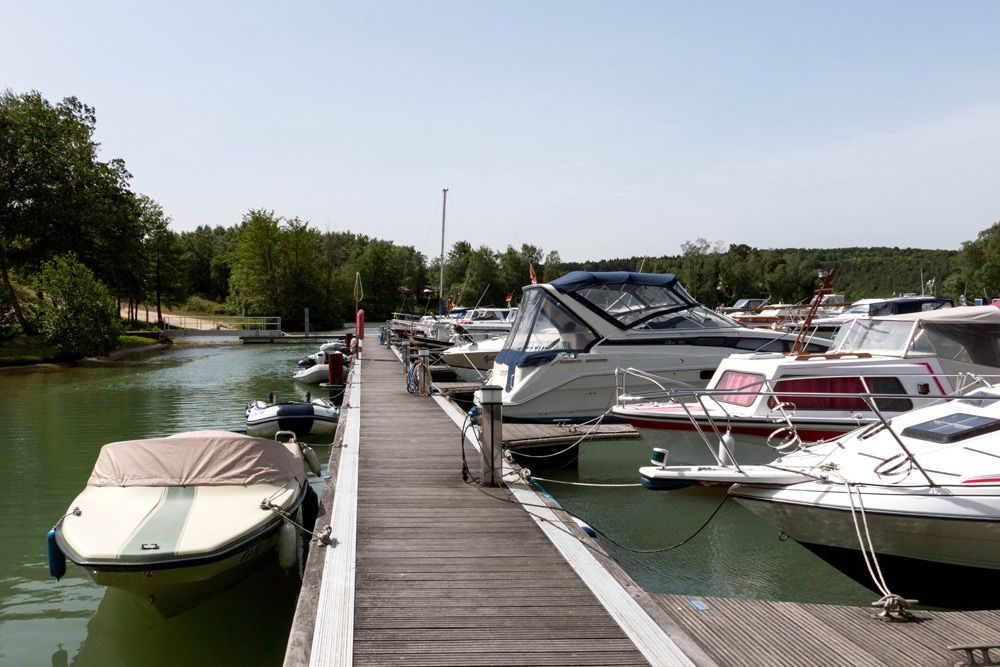 Ein hölzerner Steg mit mehreren Motorbooten und Jachten auf beiden Seiten, umgeben von Wasser und Bäumen unter einem klaren Himmel.