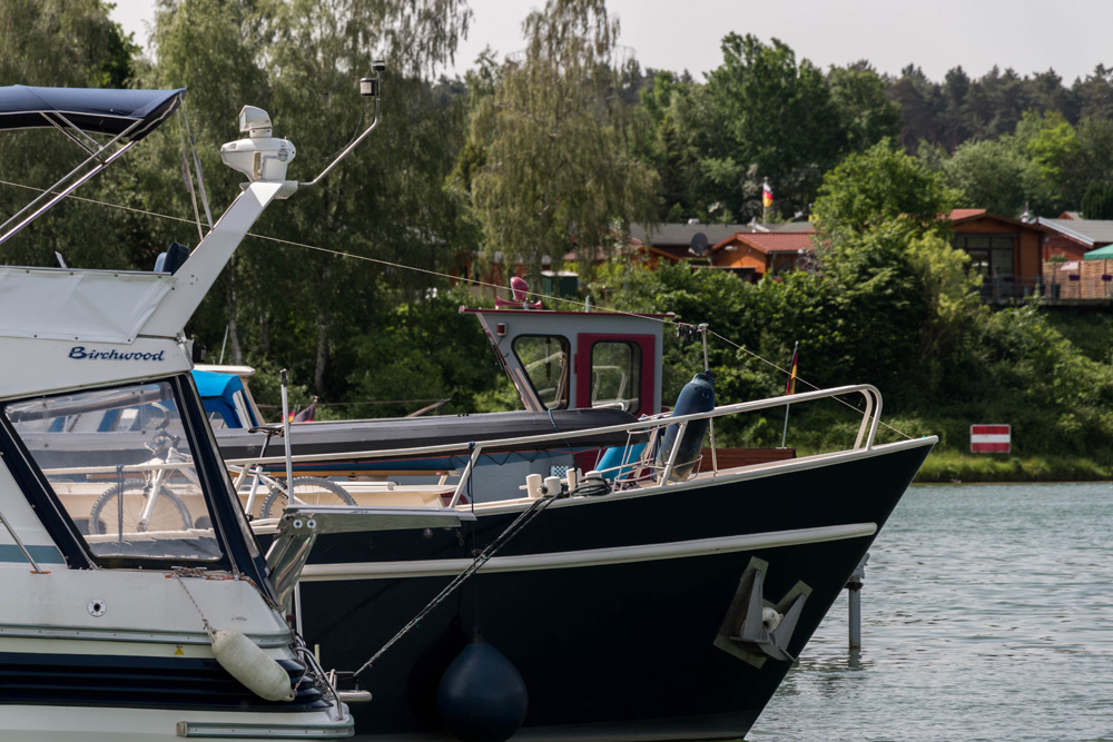 Mehrere Boote liegen an einem ruhigen Flussufer vor Anker, im Hintergrund sind Bäume und Holzhütten zu sehen.