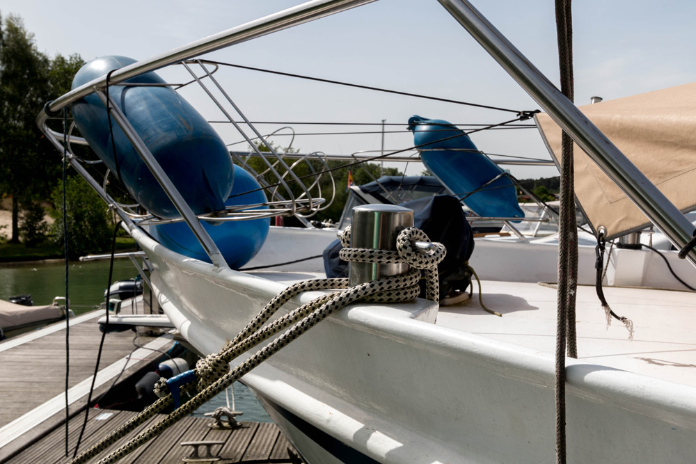 Nahaufnahme eines angedockten Bootes mit blauen Fendern und Seilen, die an einer Metallklampe auf dem Deck befestigt sind, neben einem Holzsteg.