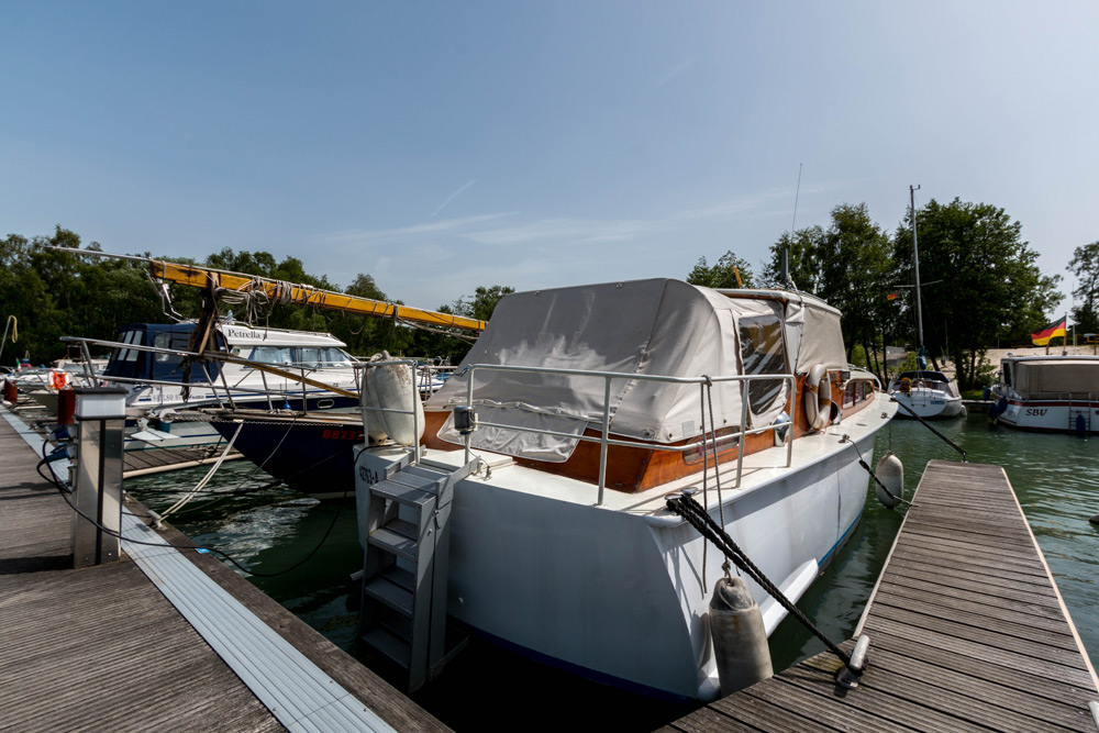 Ein weißes Motorboot mit einer überdachten Kabine liegt in einem Hafen, umgeben von anderen Booten, mit Bäumen und einem klaren Himmel im Hintergrund.