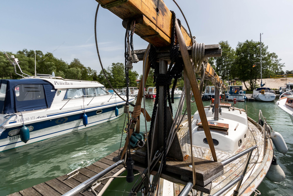 Ansicht eines angedockten Segelboots mit Takelage und Seilen im Vordergrund, umgeben von anderen Booten in einem ruhigen Yachthafen mit Bäumen im Hintergrund.