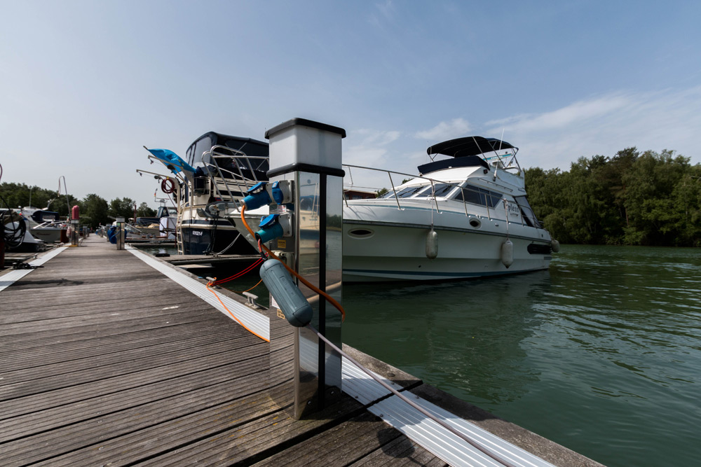Eine weiße Motoryacht liegt an einem hölzernen Steg neben anderen Booten, mit Bäumen und ruhigem Wasser im Hintergrund unter einem teilweise bewölkten Himmel.