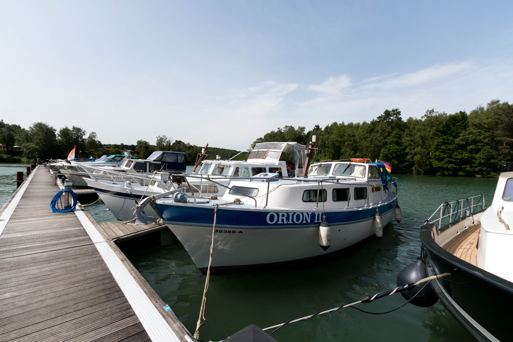 Mehrere Boote, darunter eines mit dem Namen ORION II, liegen an einem hölzernen Steg an einem See, der von Bäumen umgeben ist, unter einem teilweise bewölkten Himmel.