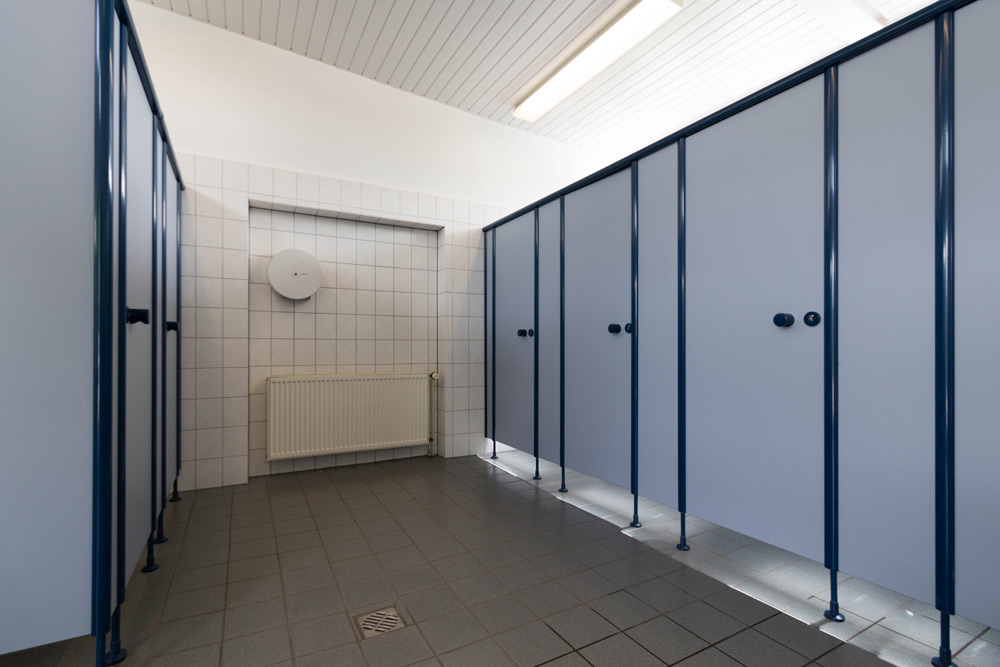 Eine öffentliche Toilette mit blauen Kabinentüren, gefliesten Wänden und Böden, einer Deckenleuchte und einem Heizkörper an der Rückwand.