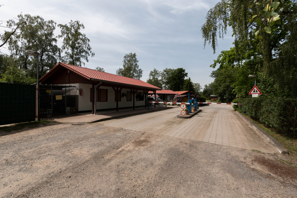 Ein kleiner Einreisekontrollpunkt mit einem Gebäude mit rotem Dach, Schranken, Verkehrsschildern und umliegenden Bäumen an einem sonnigen Tag.