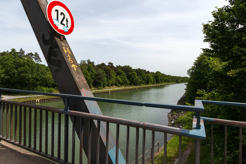 Eine Metallbrücke mit einem Schild zur Gewichtsbegrenzung von 12 Tonnen überquert einen Kanal, der auf beiden Seiten von Bäumen gesäumt ist, unter einem teilweise bewölkten Himmel.