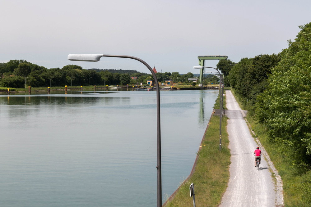 Eine Person in einer rosafarbenen Jacke fährt mit dem Fahrrad auf einem Kiesweg neben einem ruhigen Kanal, gesäumt von Bäumen und Straßenlaternen, unter einem bewölkten Himmel.