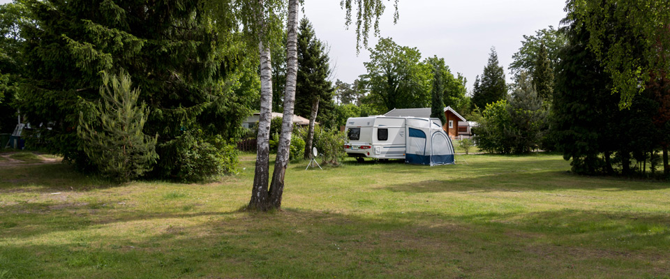 Ein weißer Wohnwagen mit Vorzelt ist auf einer von Bäumen umgebenen Wiese geparkt, im Hintergrund ist ein kleines Haus zu sehen.