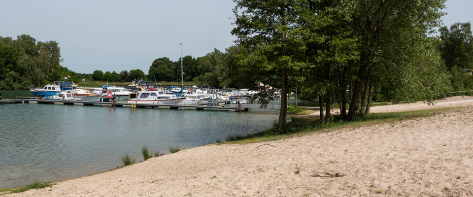 Ein Sandstrand grenzt an einen ruhigen Jachthafen mit mehreren angedockten Booten, umgeben von Bäumen und Grünflächen unter einem klaren Himmel.