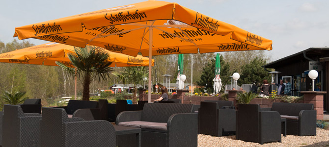 Sitzbereich im Freien mit Korbstühlen und Tischen unter großen orangefarbenen Sonnenschirmen auf einer Kiesfläche; im Hintergrund sind ein Gebäude und Bäume zu sehen.