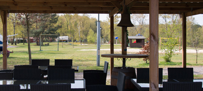Überdachter Sitzbereich im Freien mit leeren schwarzen Stühlen und Tischen, mit Blick auf einen grasbewachsenen Park mit Bäumen, Spielgeräten und einem Volleyballfeld im Hintergrund.