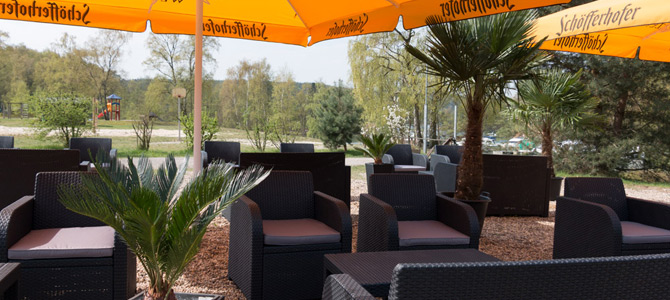 Sitzbereich im Freien mit schwarzen Korbstühlen und Tischen unter großen gelben Sonnenschirmen, umgeben von Bäumen und kleinen Palmenpflanzen.