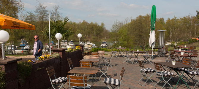 Außencafé mit leeren Tischen und Stühlen, ein Mann steht an einer Wand, Blumen in Pflanzkübeln, Bäume und Boote sind im Hintergrund unter einem teilweise bewölkten Himmel zu sehen.