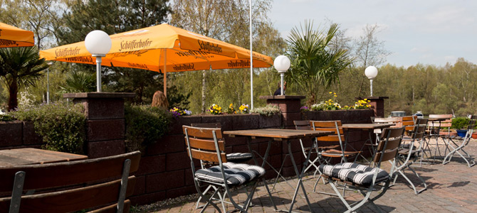 Außenterrasse mit Holztischen und -stühlen, gestreiften Kissen, gelben Sonnenschirmen und Topfpflanzen an einem sonnigen Tag.