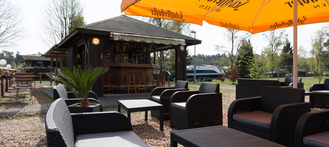 Sitzbereich im Freien mit Korbstühlen und Tischen unter großen orangefarbenen Sonnenschirmen in der Nähe einer kleinen Holzbar, umgeben von Bäumen und Grün.