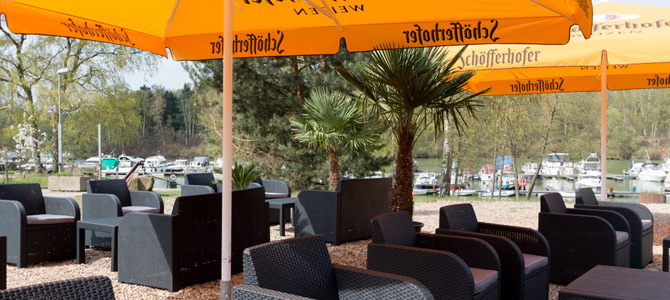 Sitzbereich im Freien mit schwarzen Korbstühlen und Tischen unter großen orangefarbenen Schöfferhofer-Sonnenschirmen, mit Blick auf einen Yachthafen mit Booten und umgeben von Bäumen.