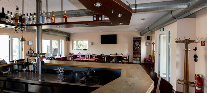 Modernes Barinterieur mit Holztheke, Spülbecken, Flaschenauslage, Sitzgelegenheiten, Wandfernseher, Dartscheibe und Garderobe.