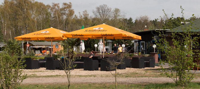 Sitzbereich im Freien mit schwarzen Stühlen und Tischen unter großen gelben Schöfferhofer-Schirmen, umgeben von kleinen Bäumen und Pflanzen in der Nähe eines Gebäudes.