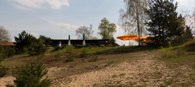 Eine abschüssige Rasenfläche mit vereinzelten Bäumen, die zu einem Gebäude mit geschlossenen Schirmen und einem großen offenen orangefarbenen Regenschirm im Freien führt.