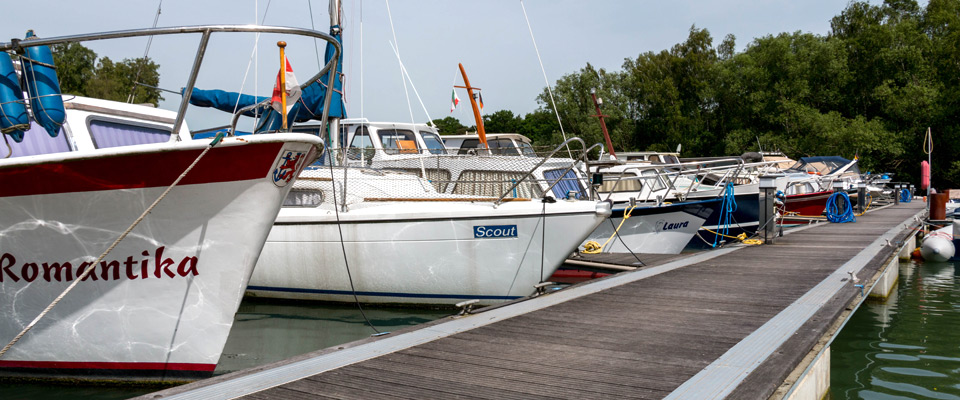 Mehrere Boote liegen an einem Holzsteg an einem ruhigen Gewässer, im Hintergrund sind Bäume zu sehen.